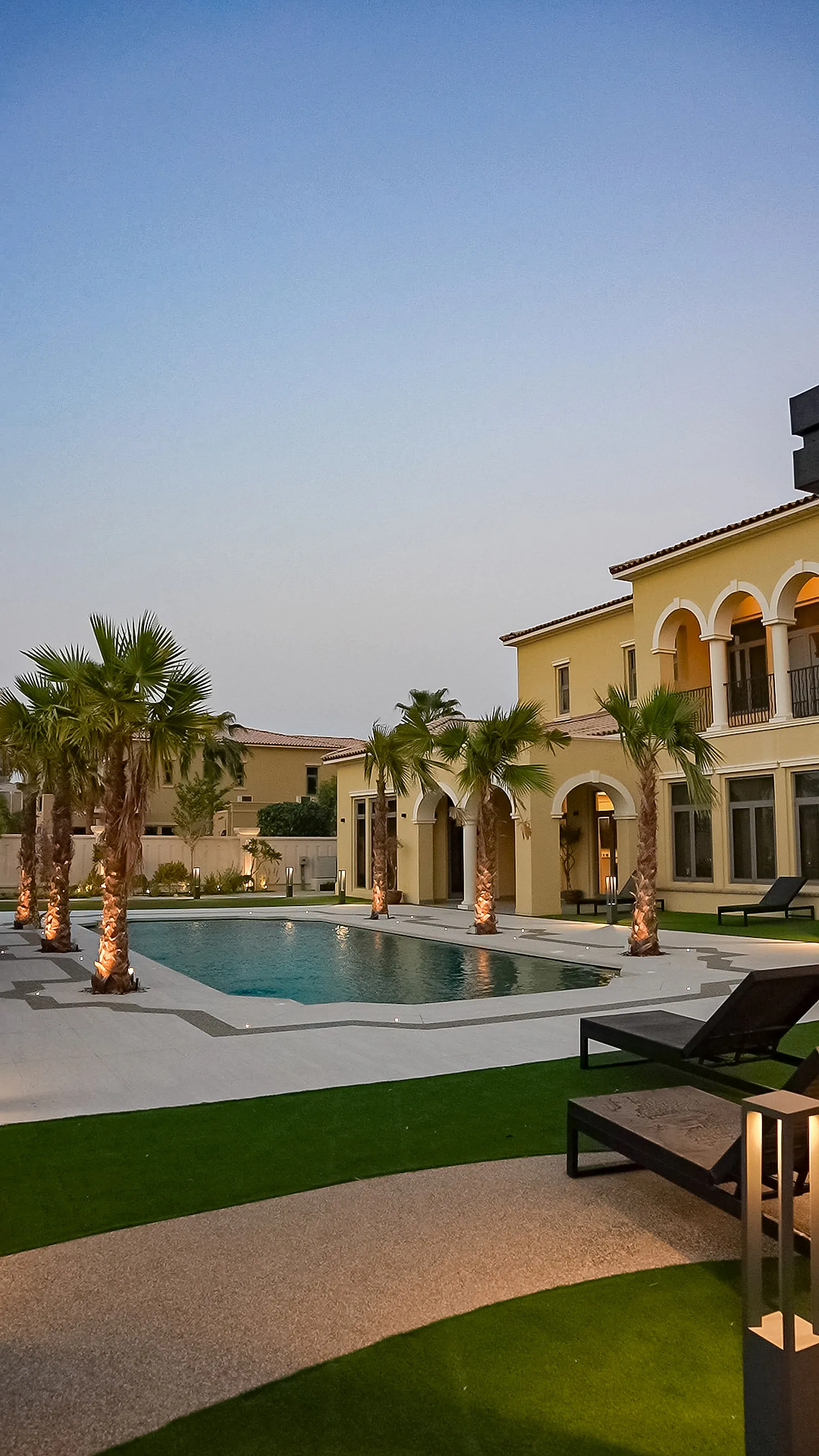 The classical villa beyond the pool at dusk, with uplit palms and low-level fittings along the lawn