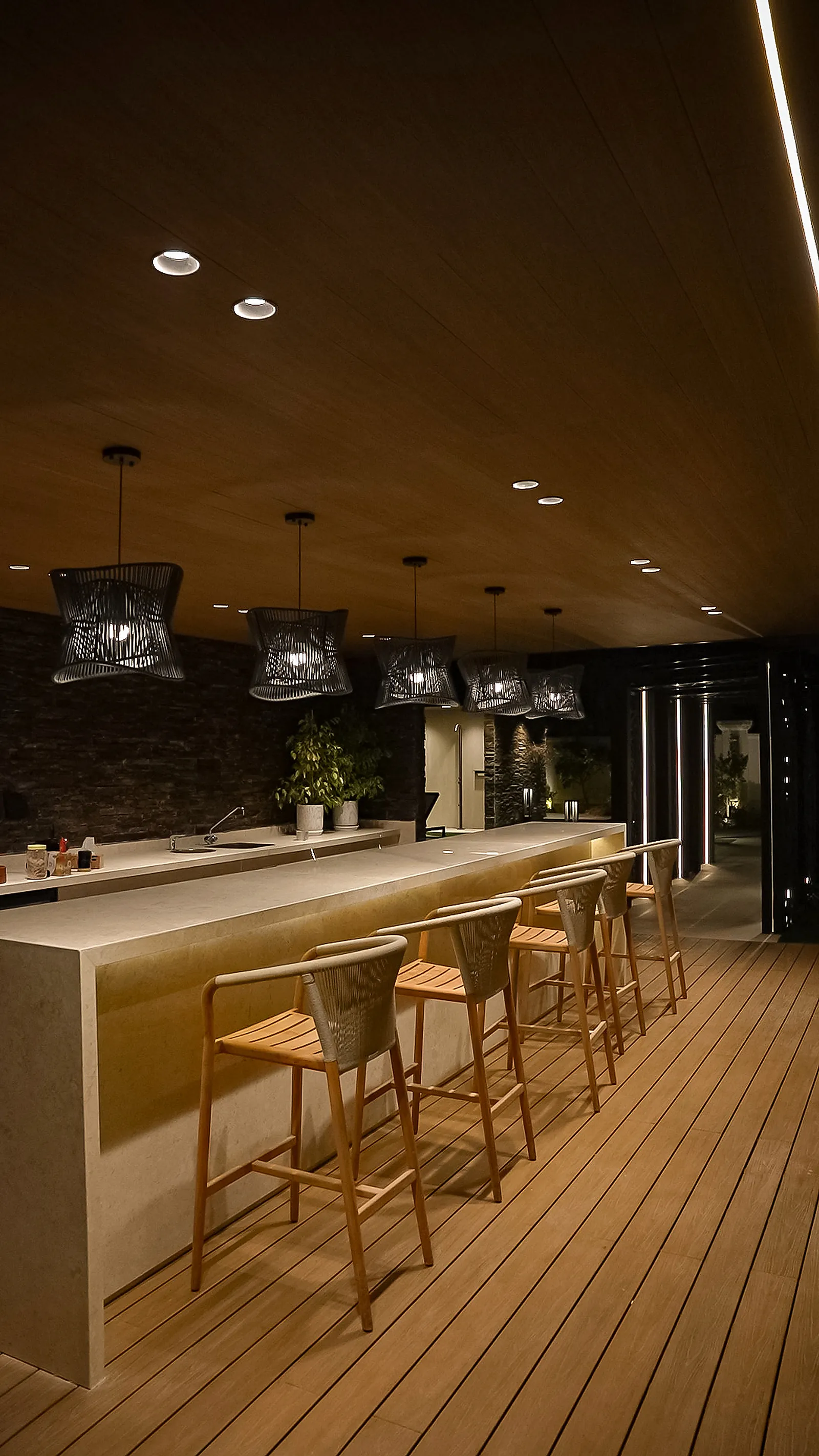 The outdoor bar counter at night, lit by pendants, soffit downlights and a strip concealed beneath the counter overhang