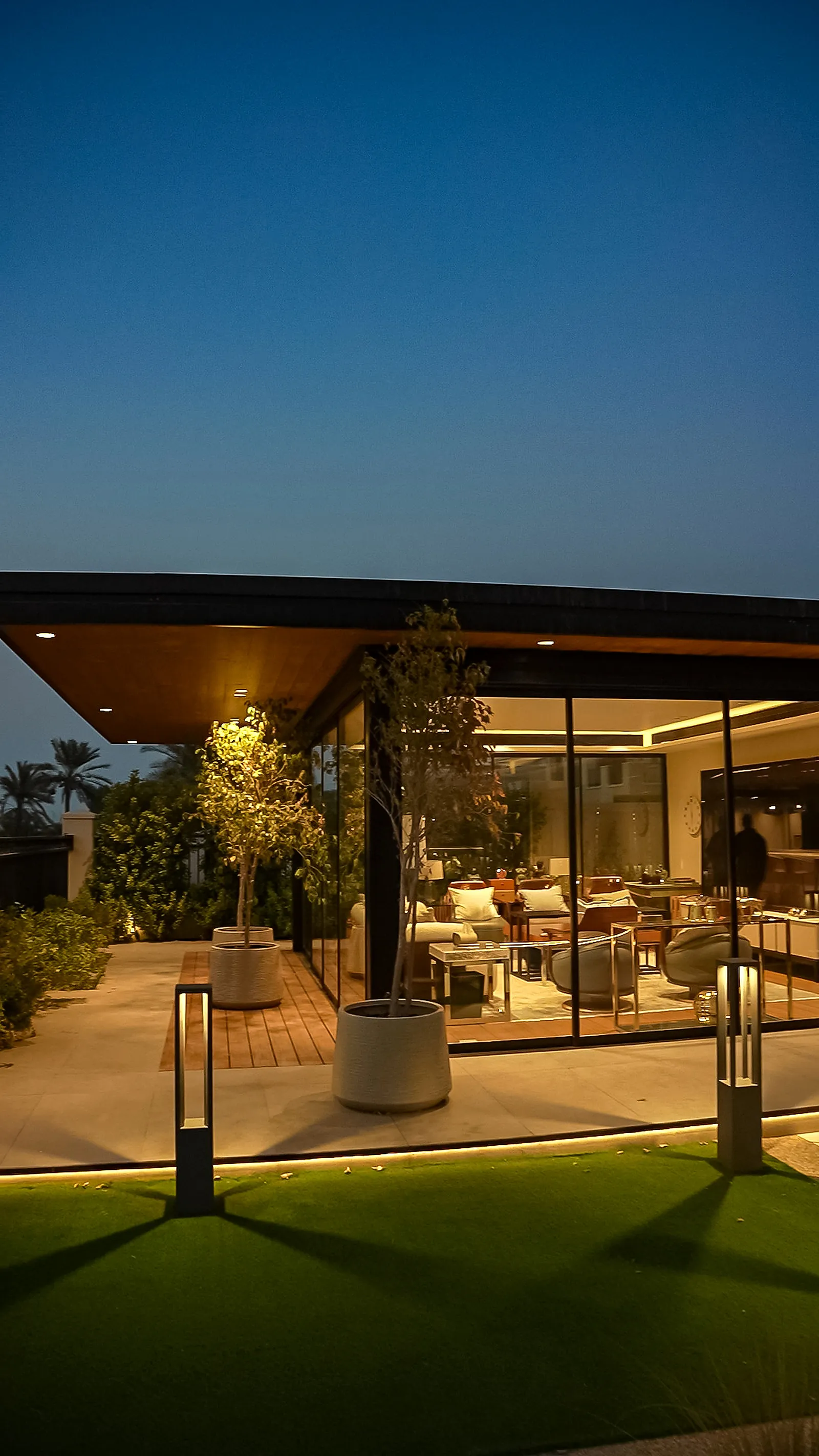 The glazed outdoor lounge at dusk, with bollards on the lawn and warm light carrying out from inside