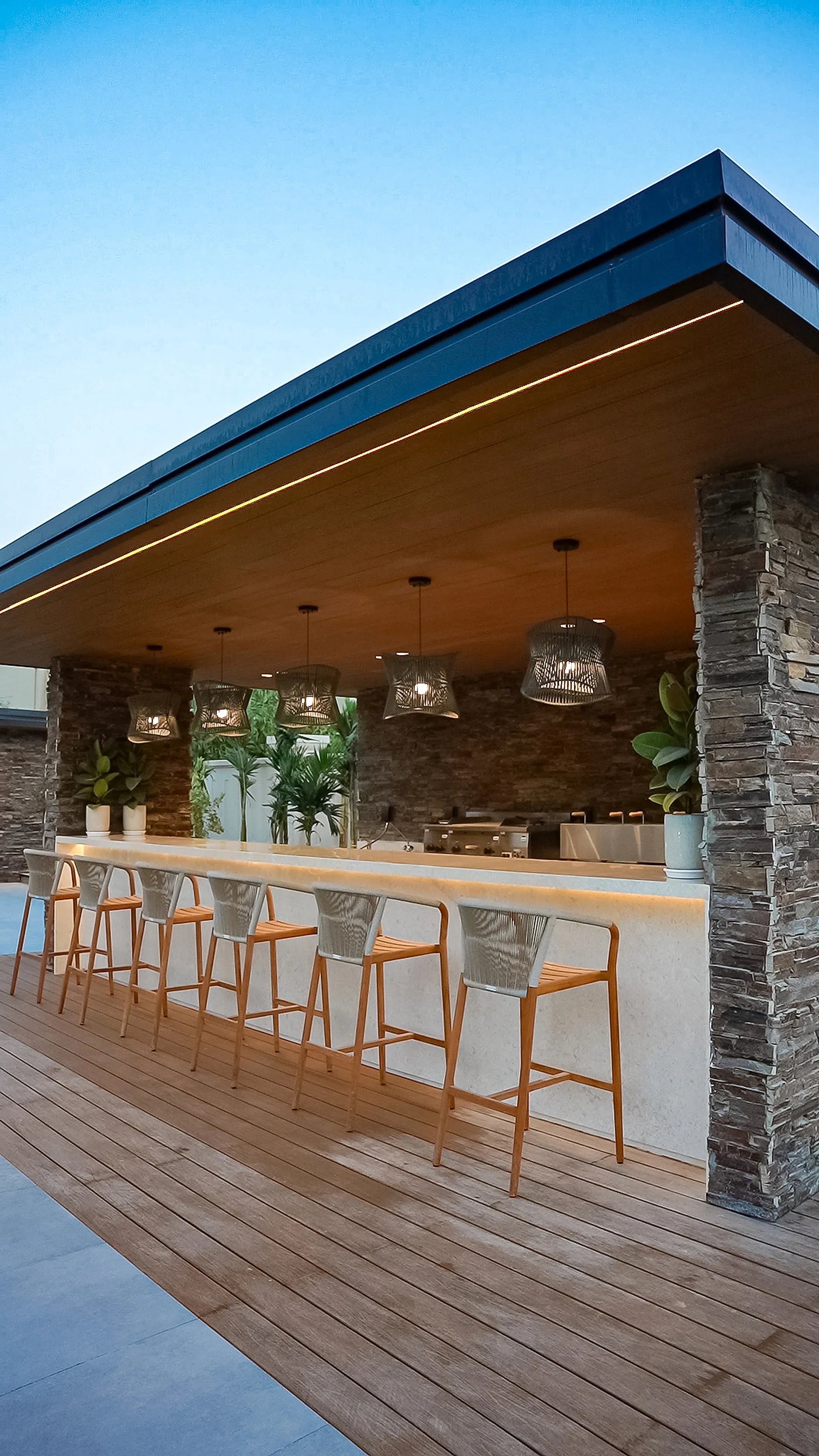 The bar pavilion at the Abu Dhabi villa at dusk, with a concealed strip at the roof edge, downlights in the timber soffit and woven pendants over the counter