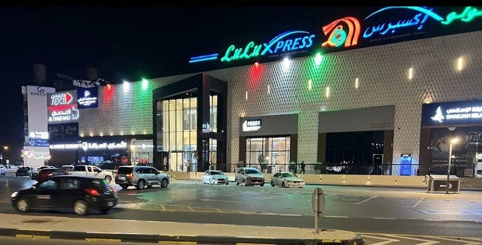 Al Dhaid Mall at night, its textured facade washed in colour with illuminated tenant signage above the entrance canopy