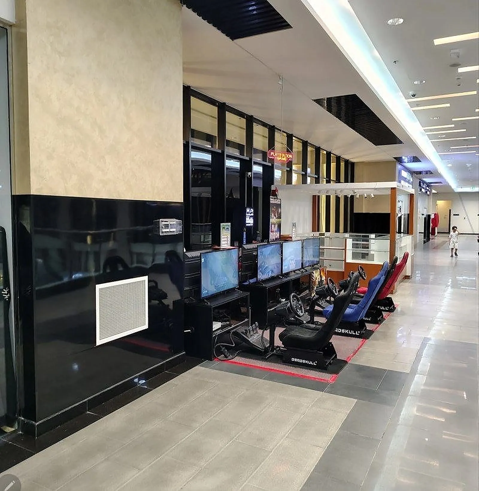An entertainment corridor inside Al Dhaid Mall, with recessed downlights and a linear cove in the mall ceiling and a brighter tenant unit alongside