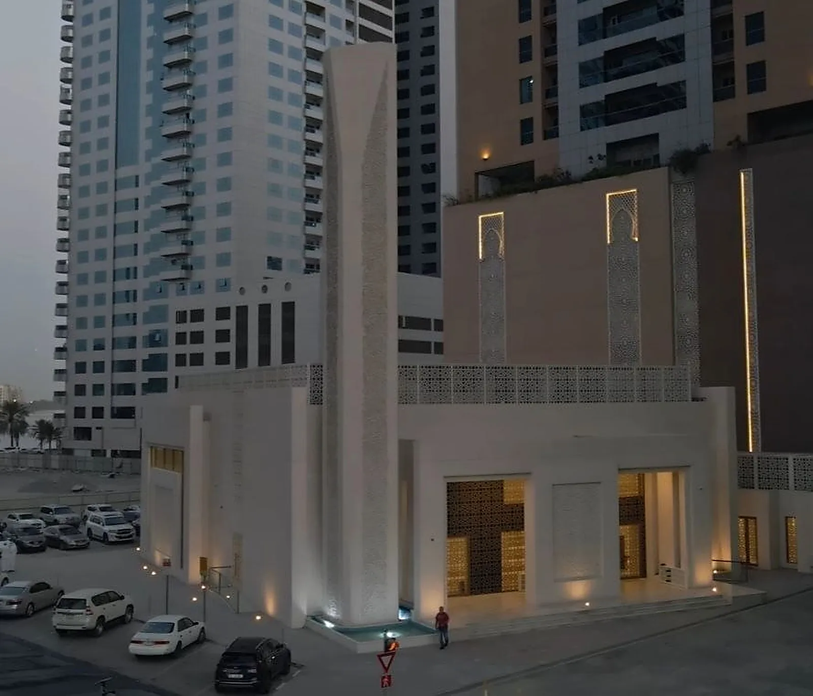 Al Rajaa Mosque in Sharjah at dusk, its minaret uplit from the ground, perforated geometric screens glowing along the parapet and warm light coming through the entrance screens