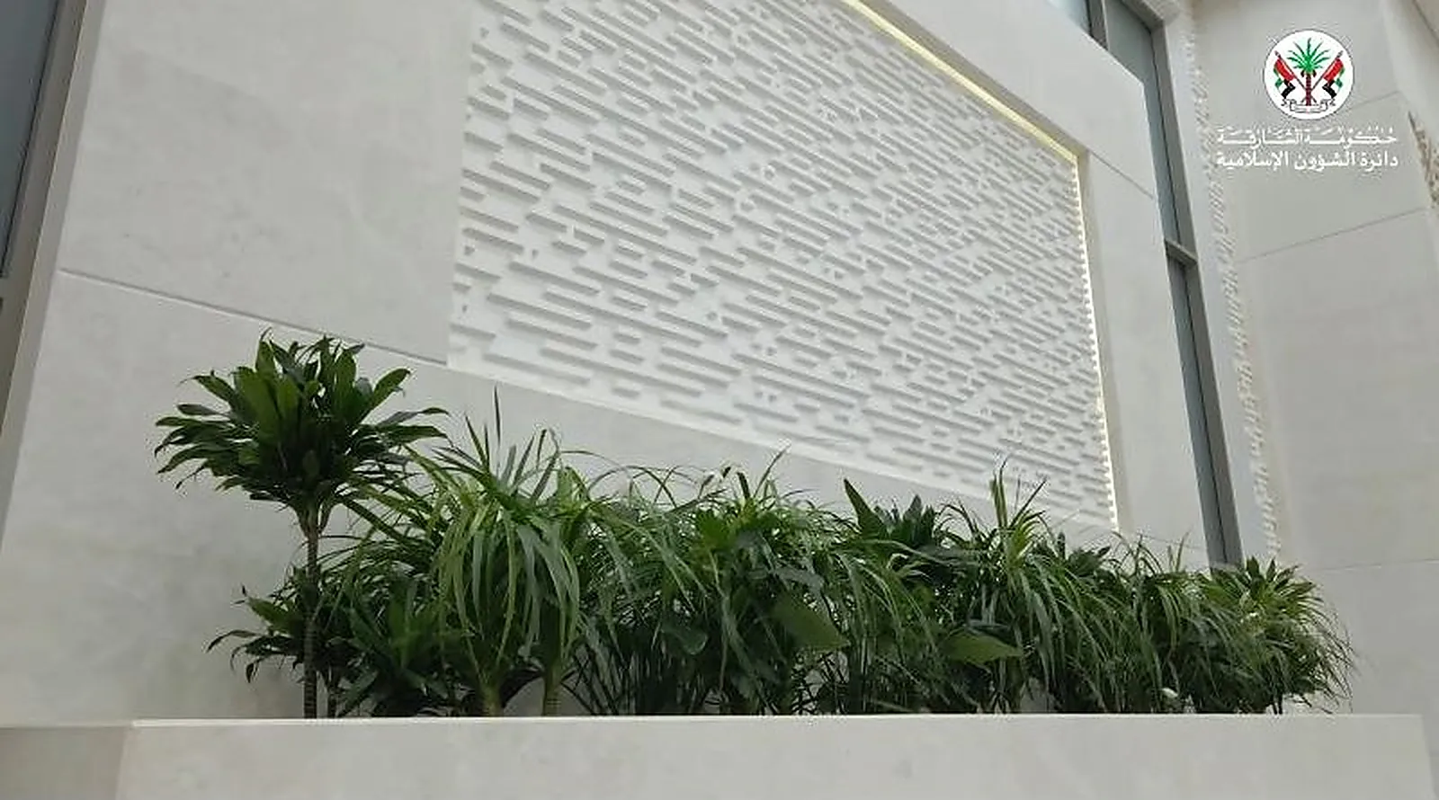 A carved calligraphic panel at Al Rajaa Mosque grazed from a concealed source along its top edge, with planting below it