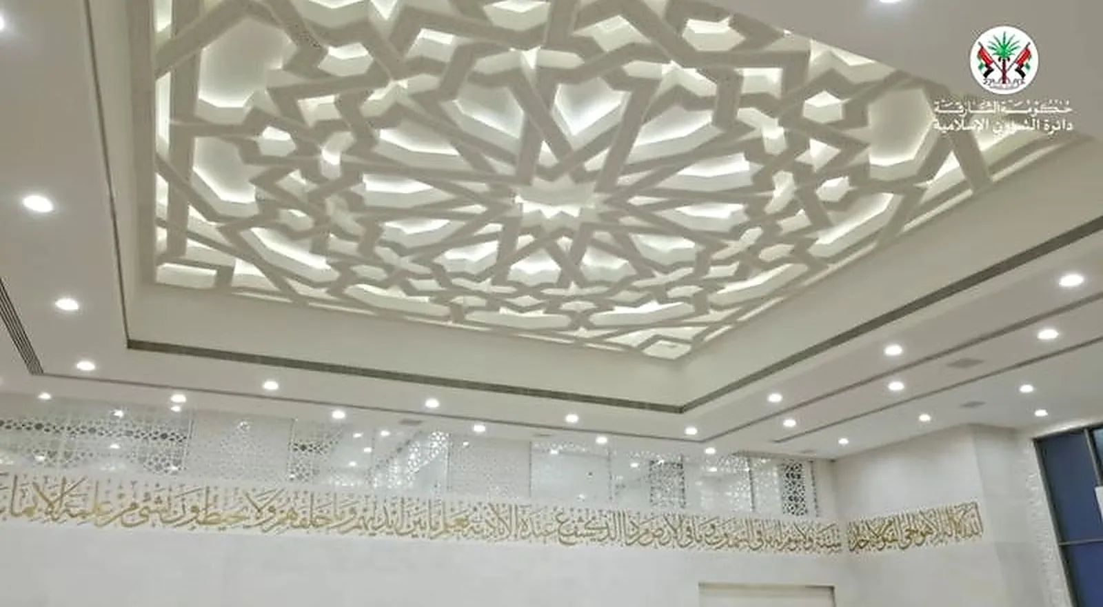 The prayer hall ceiling at Al Rajaa Mosque, a carved geometric panel backlit inside a deep coffer with downlights in the flat soffit around it and a calligraphic frieze on the wall below