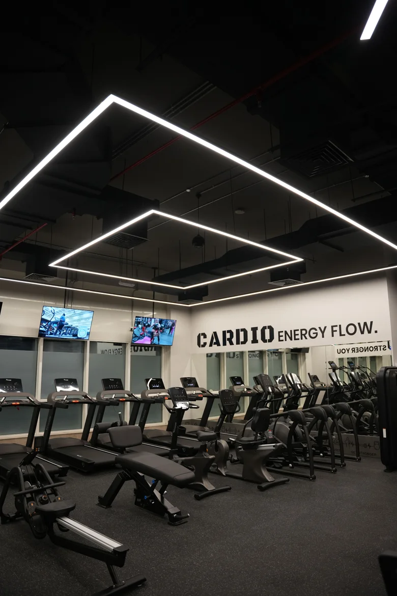 The cardio floor at Al-Serh Gym, a row of treadmills and benches beneath a closed rectangle of suspended linear light on the dark soffit