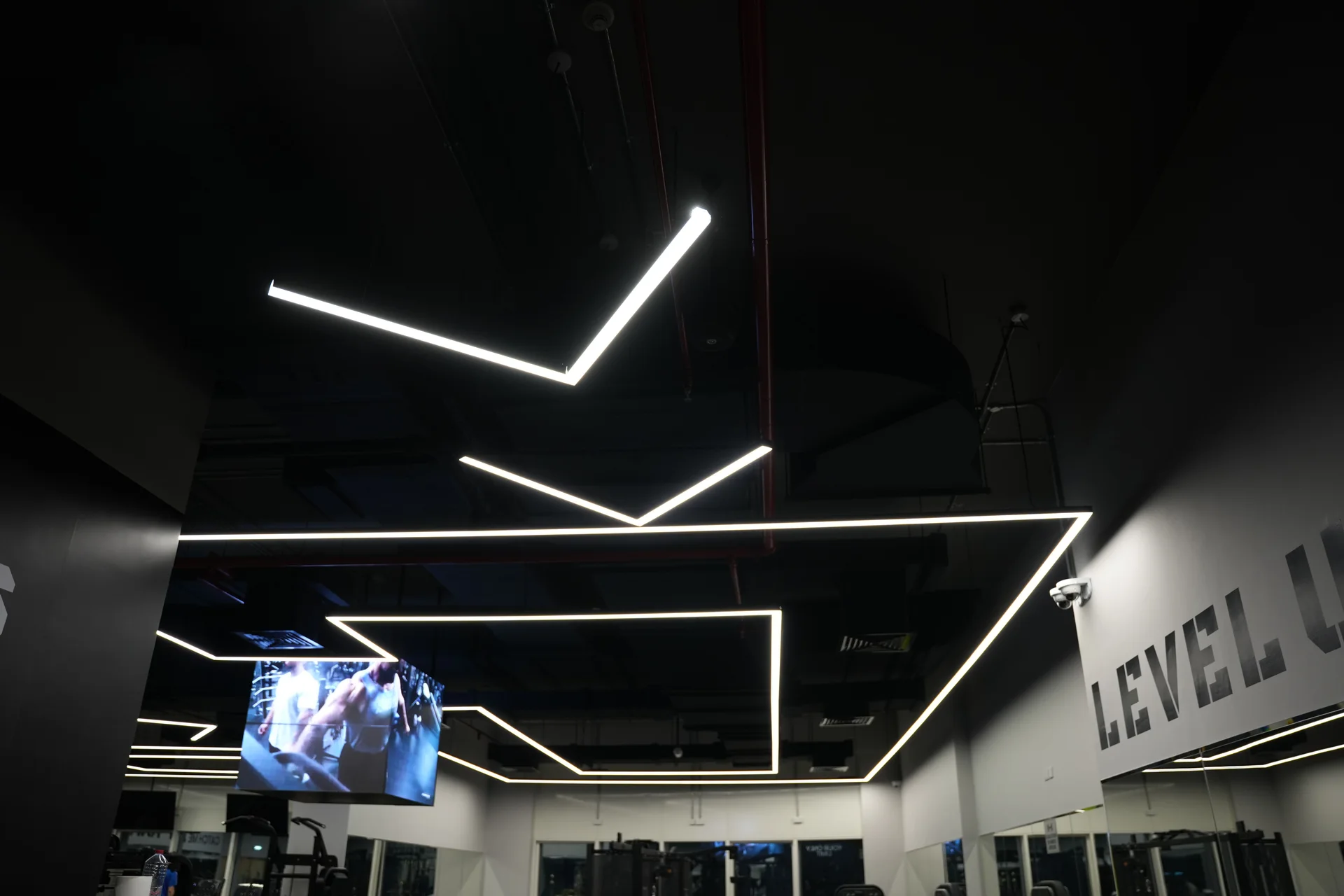 The training floor at Al-Serh Gym in Sharjah, its dark exposed ceiling carrying suspended linear profile set out as angular figures above the equipment