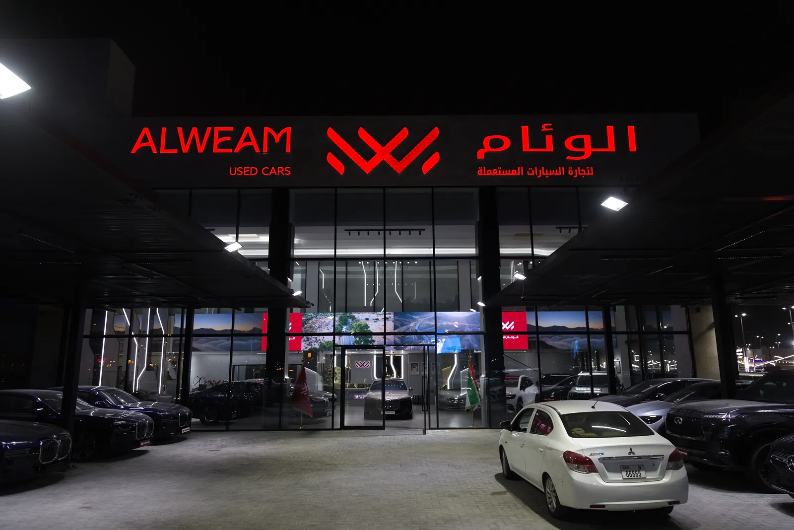 The ALWEAM Used Cars entrance at night, its red channel-letter signage lit above a glazed frontage with cars parked either side