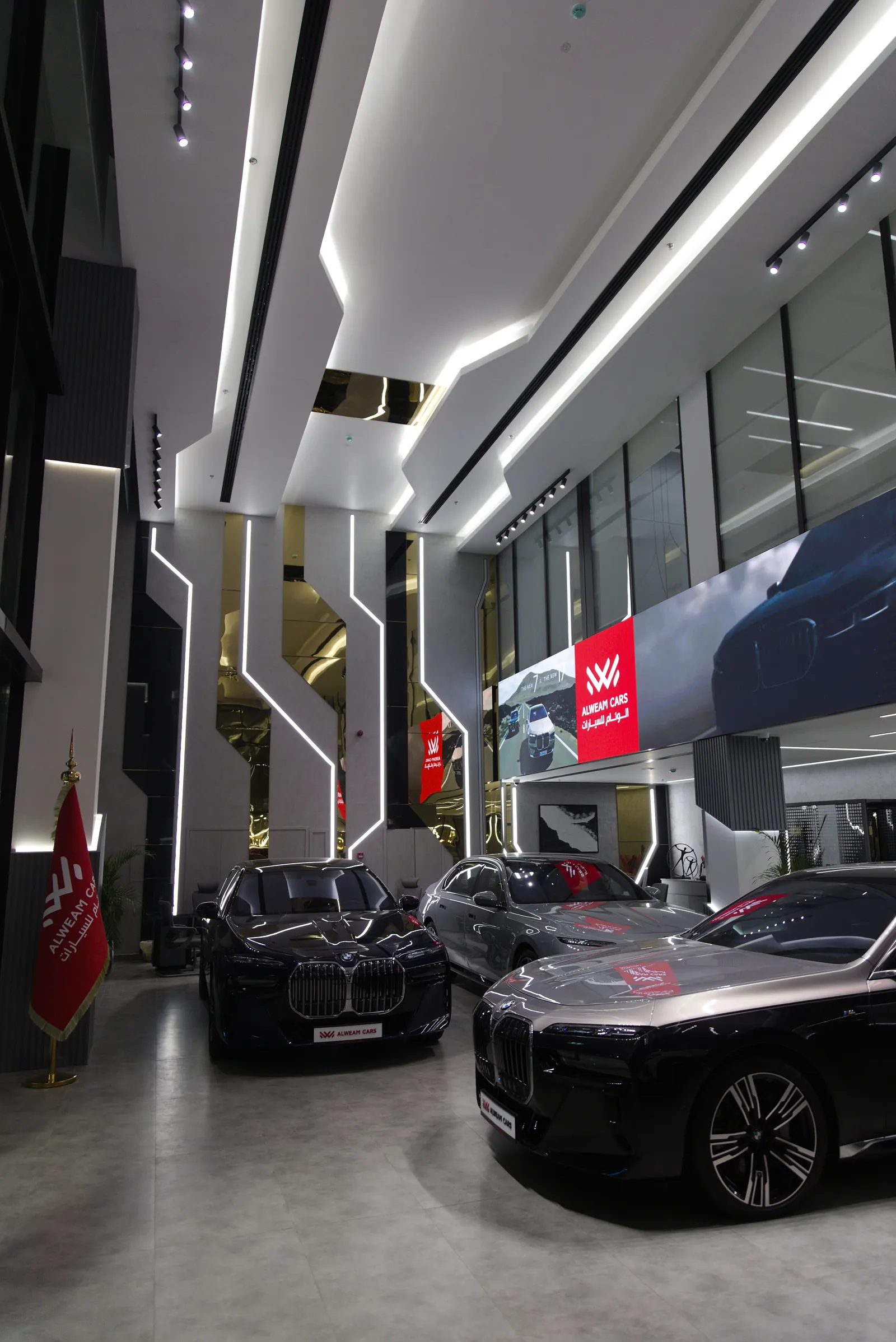 The vehicle display floor at Al Weam, with recessed linear runs in a coffered ceiling and a large LED screen behind the cars