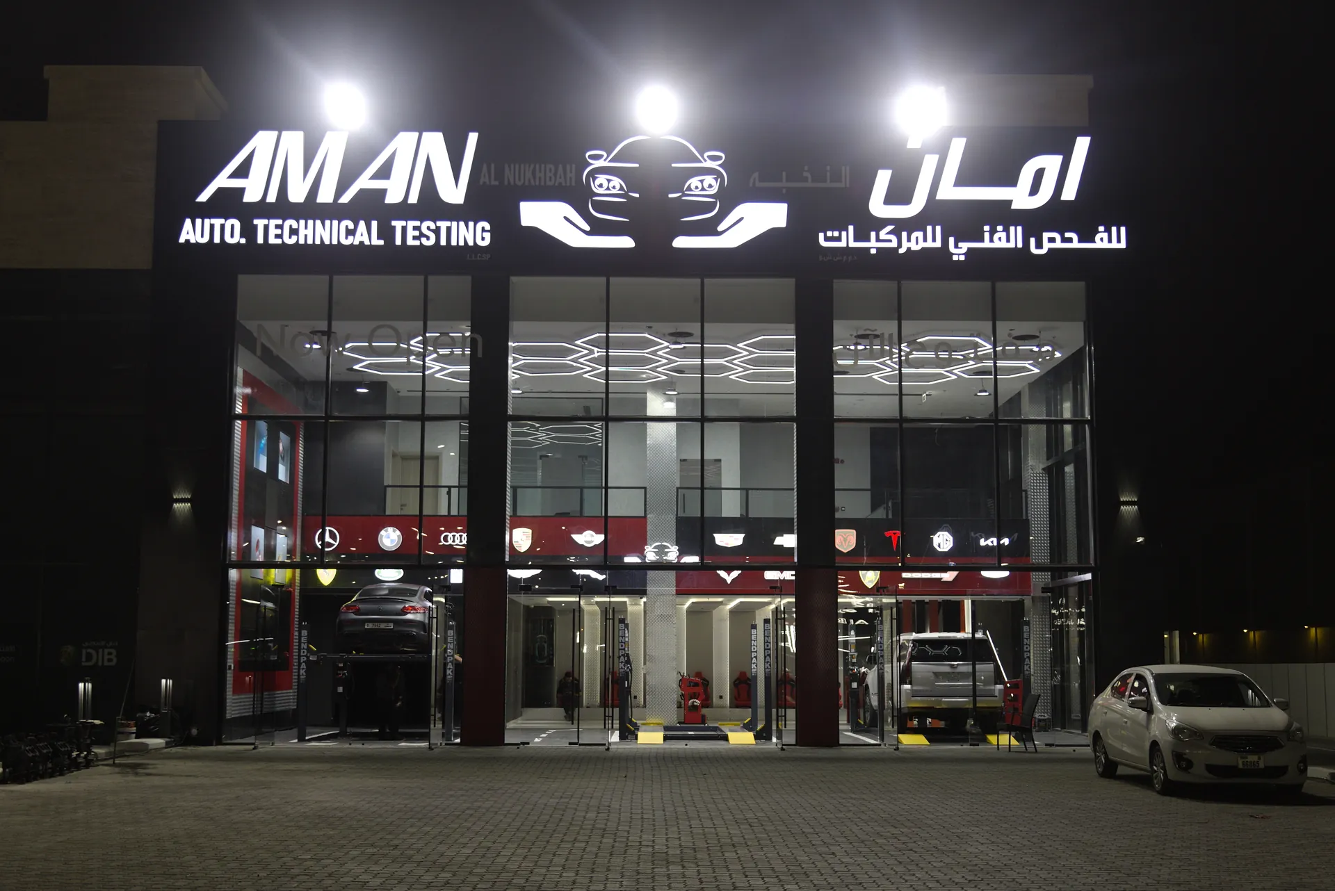 Al Weam and Al Aman in Sharjah at night, the AMAN Auto Technical Testing signage lit above a glazed frontage with the hexagonal ceiling visible inside