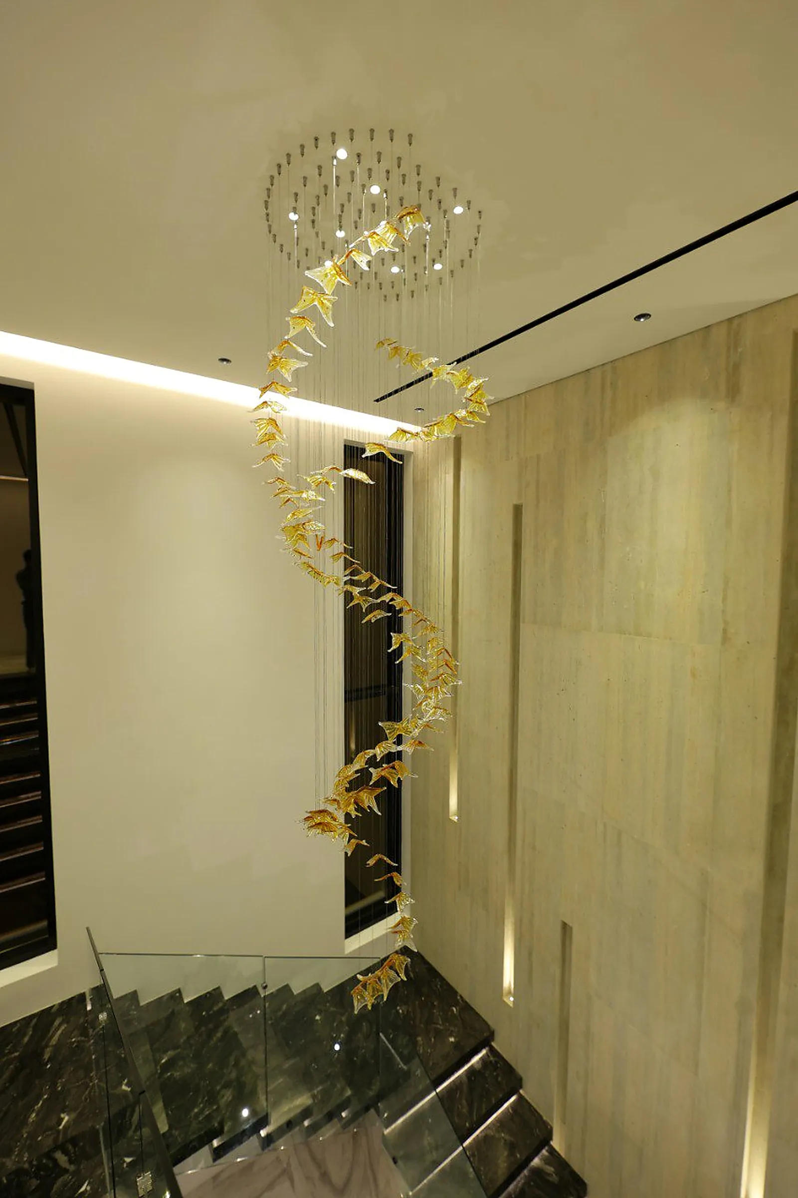 A spiral of amber glass leaves on clear cables dropping the full height of the Alkhafaji villa stairwell