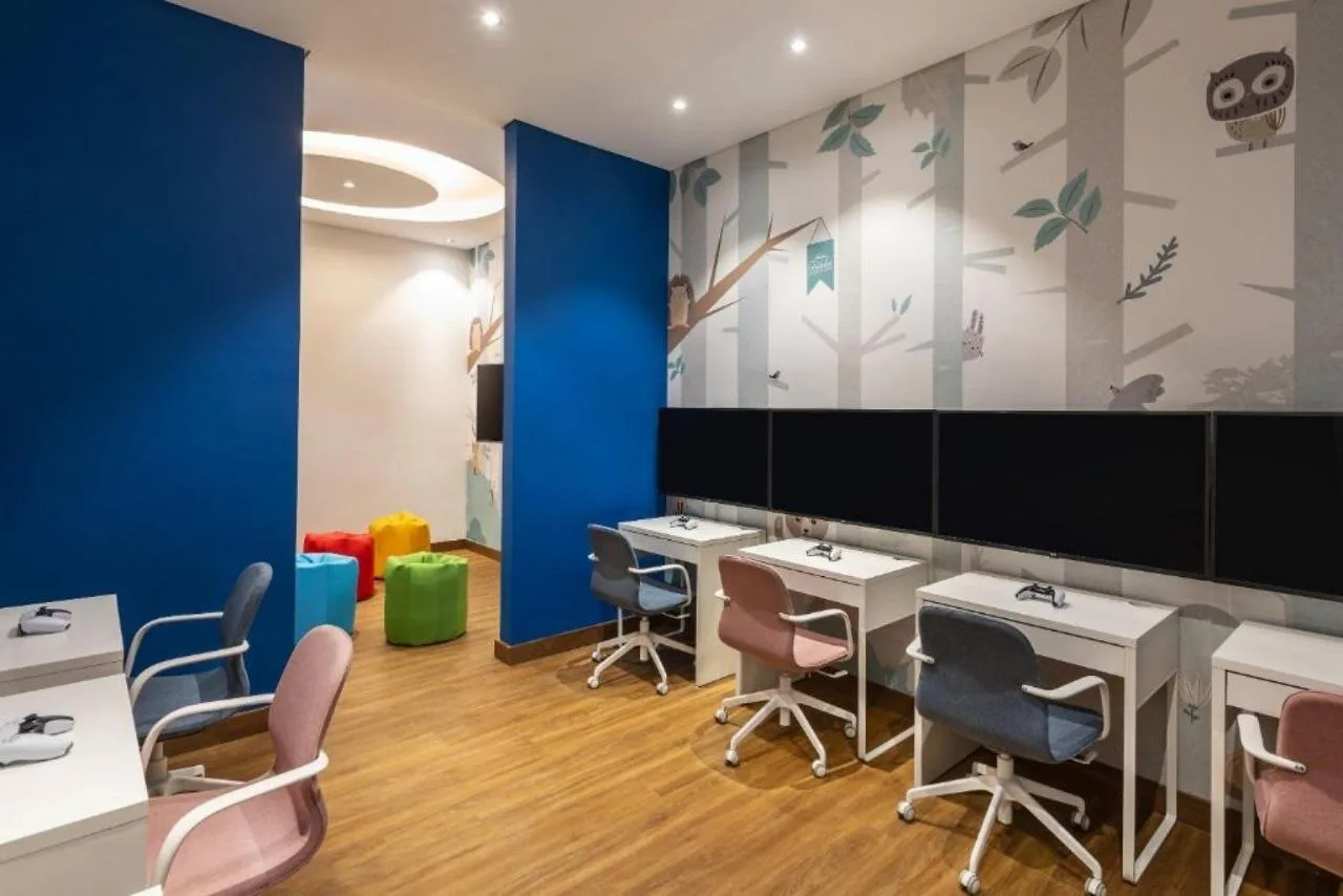 A children&rsquo;s gaming room at Aykon City with screens along one wall, small desks and chairs, and a forest mural