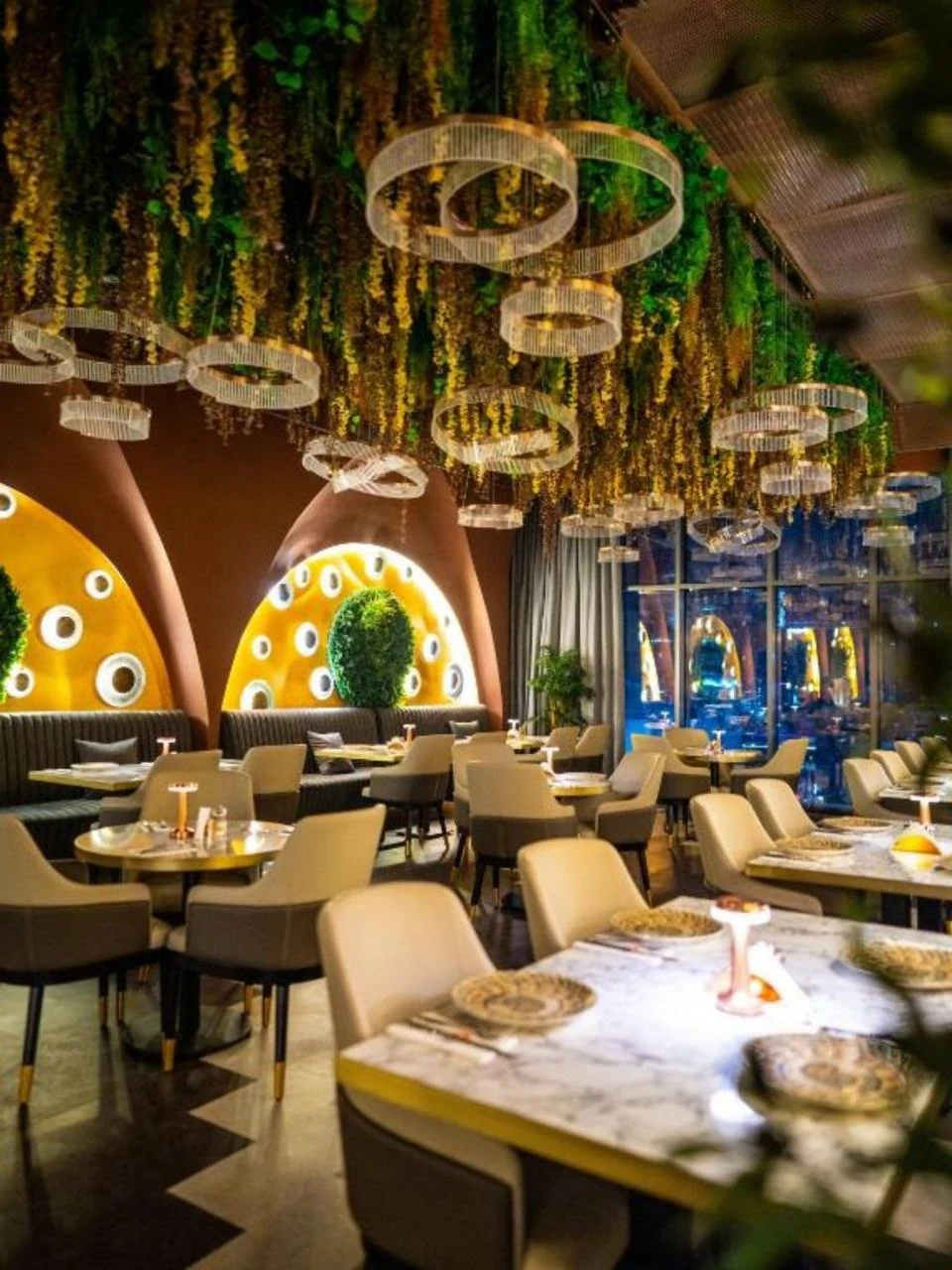 A restaurant at Aykon City lit by crystal ring pendants under a planted ceiling, with backlit arched niches behind the banquettes