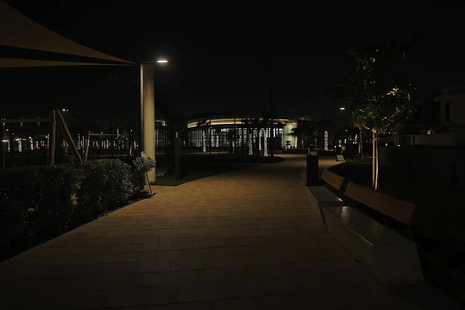 A paved walkway at night in the Dubai South residential district, lit by pole-top luminaires, with benches, clipped hedges and an uplit tree