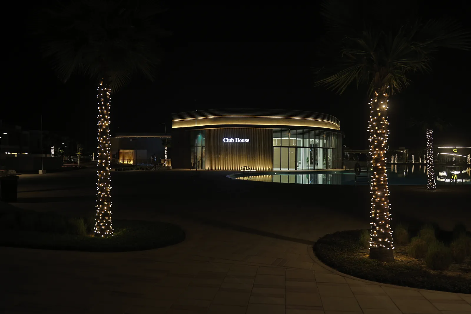The clubhouse at Dubai South seen across the plaza at night, its curved fascia lit as a horizontal band, framed by two string-lit palms
