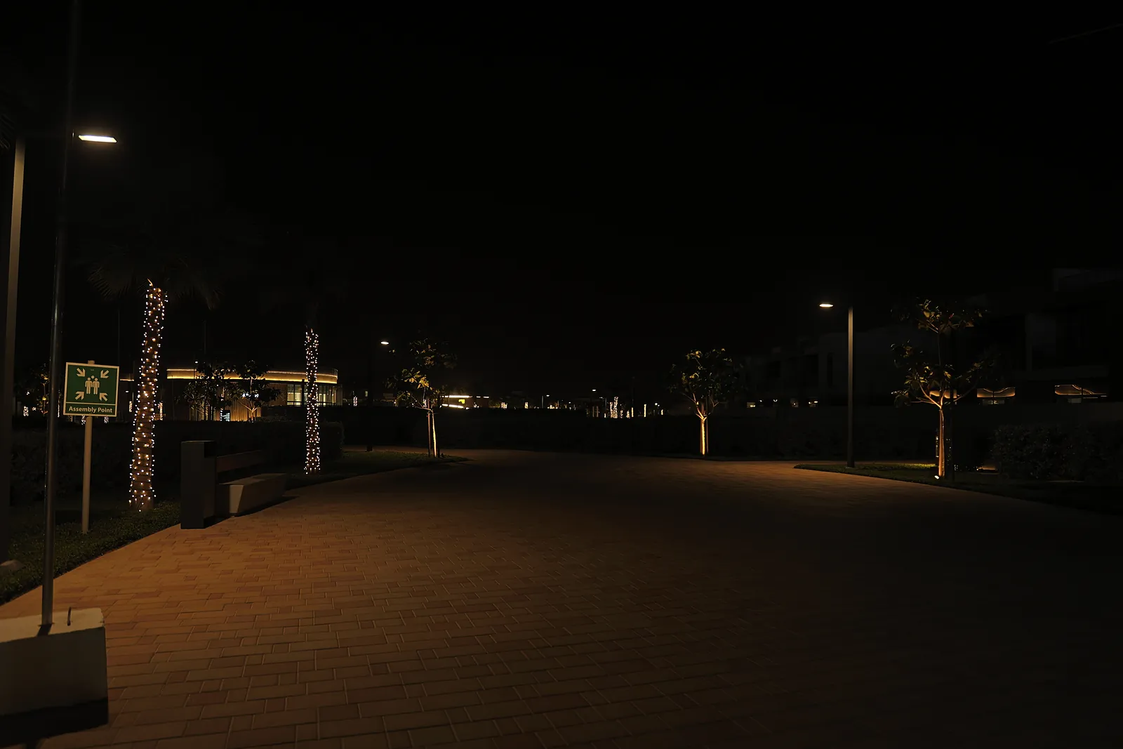 A walkway at Dubai South at night beside an assembly point sign, with a bench, a litter bin and pole-top lighting