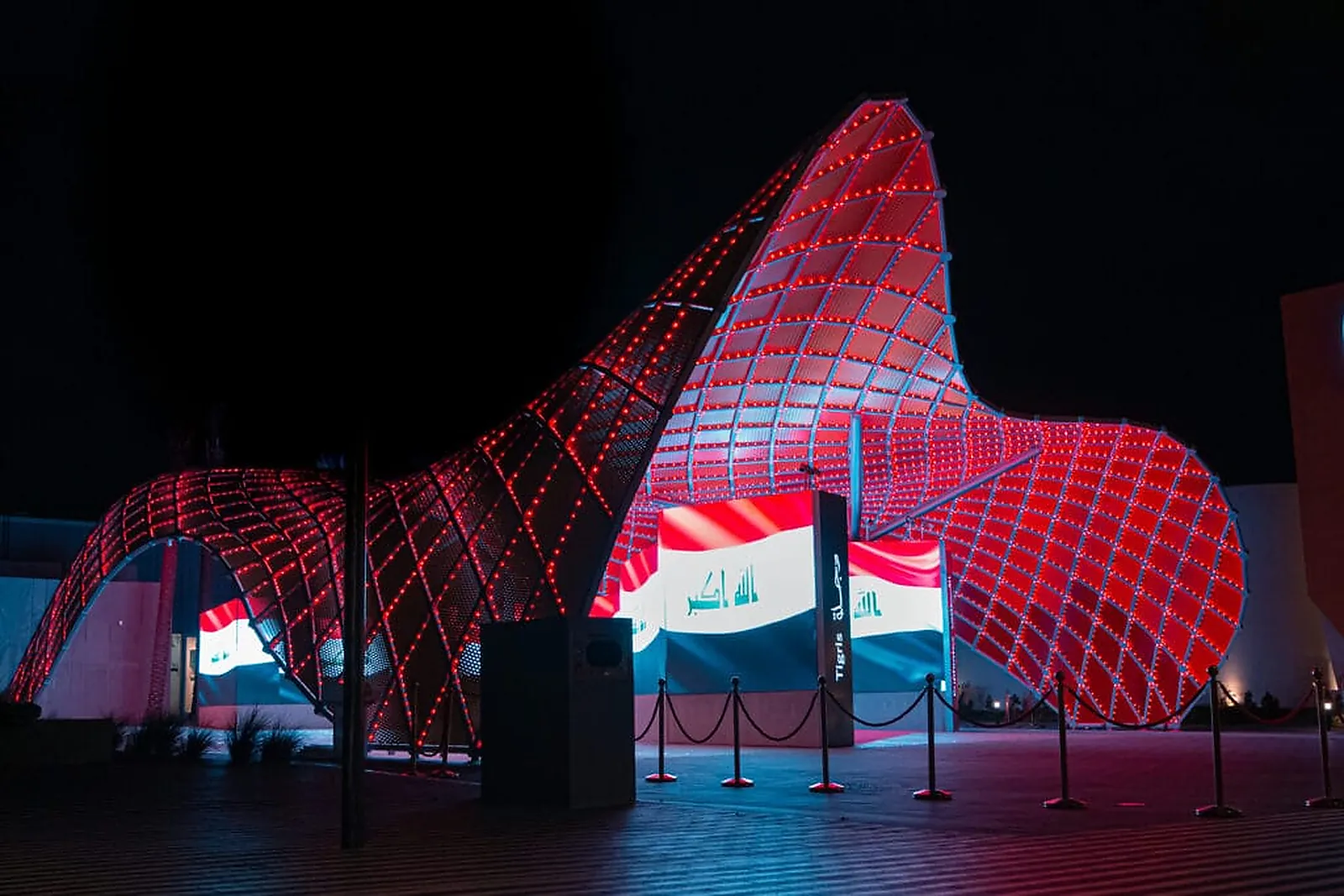 The pavilion gridshell in deep red, with the Iraqi flag lit alongside it