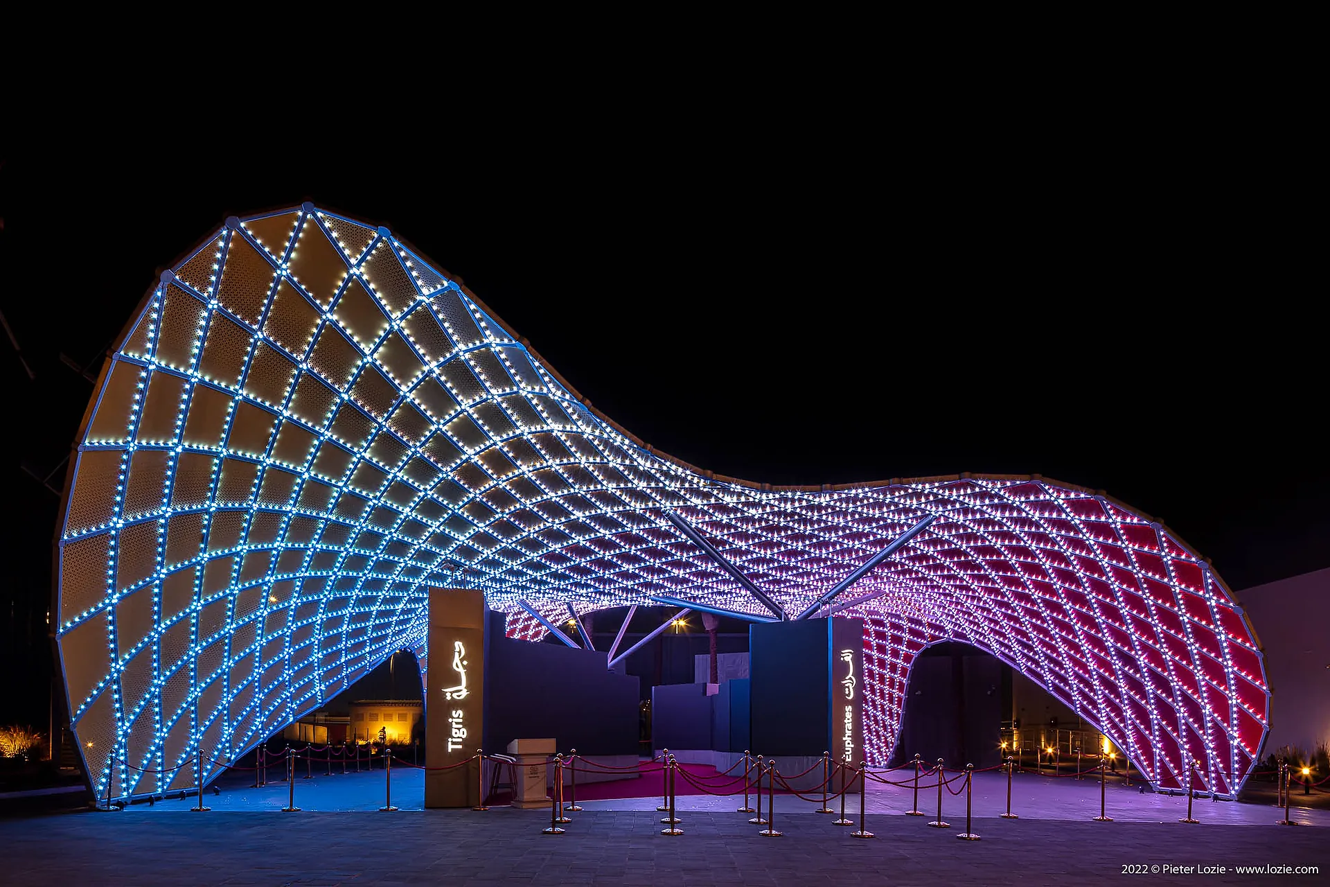 The Iraq Pavilion at Expo 2020 Dubai at night, its curved timber gridshell lit blue on one flank and red on the other