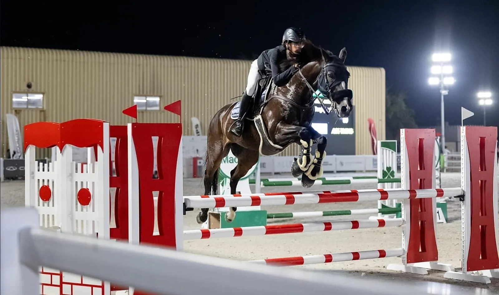 A horse and rider clearing an oxer in the floodlit outdoor arena at Mandara Equestrian Club